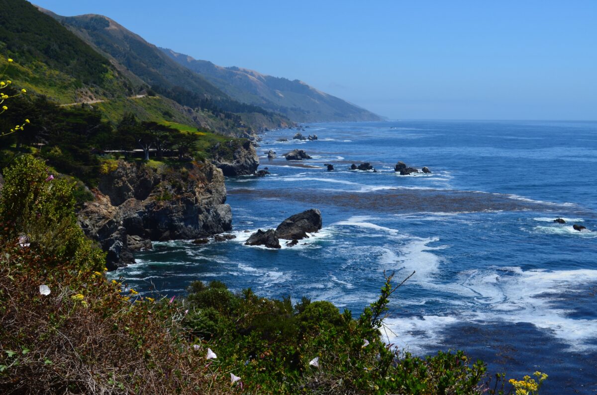 blick von oben auf ein felsiges küstenstück am highway 1 in californien, man sieht gischt, aus dem wasser ragen schwarze felsstücke