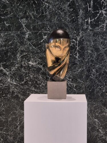 Skulptur von Brancusi, eine kleiner Torso mit rundem Kopf und verschränkten Armen, aus Bronze vor einer Marmorwand